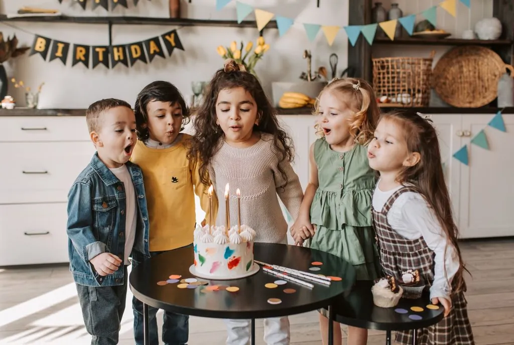 A stock photo of five children blowing out birthday candles in a kitchen